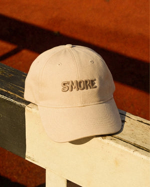 A beige baseball cap with 'SMORE' embroidered on the front, resting on a weathered wooden railing with a warm orange background.